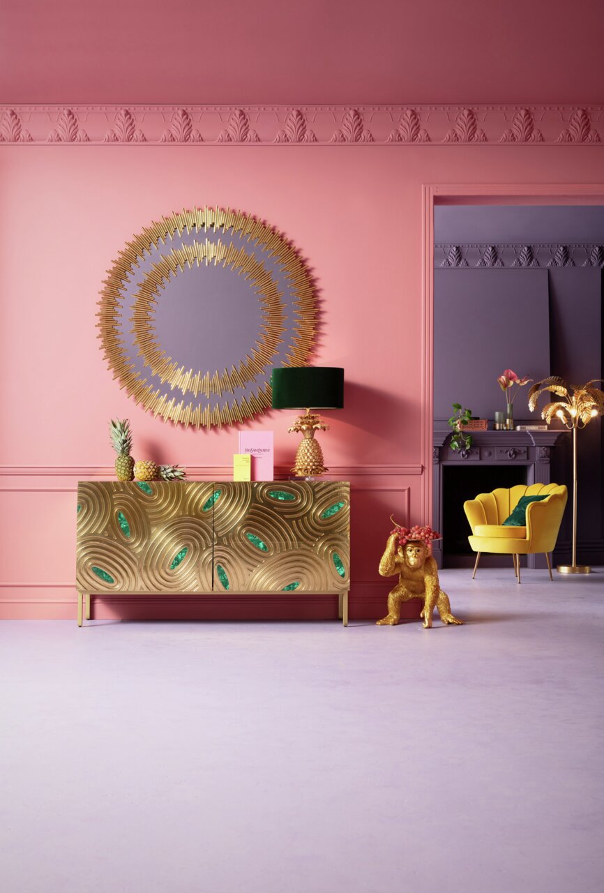 Goldene Deko-Figur eines Affen als Butler, der eine Schale mit Früchten trägt, in einem stilvoll eingerichteten Raum mit rosa Wänden und goldenen Akzenten. Perspektive: Frontalansicht.