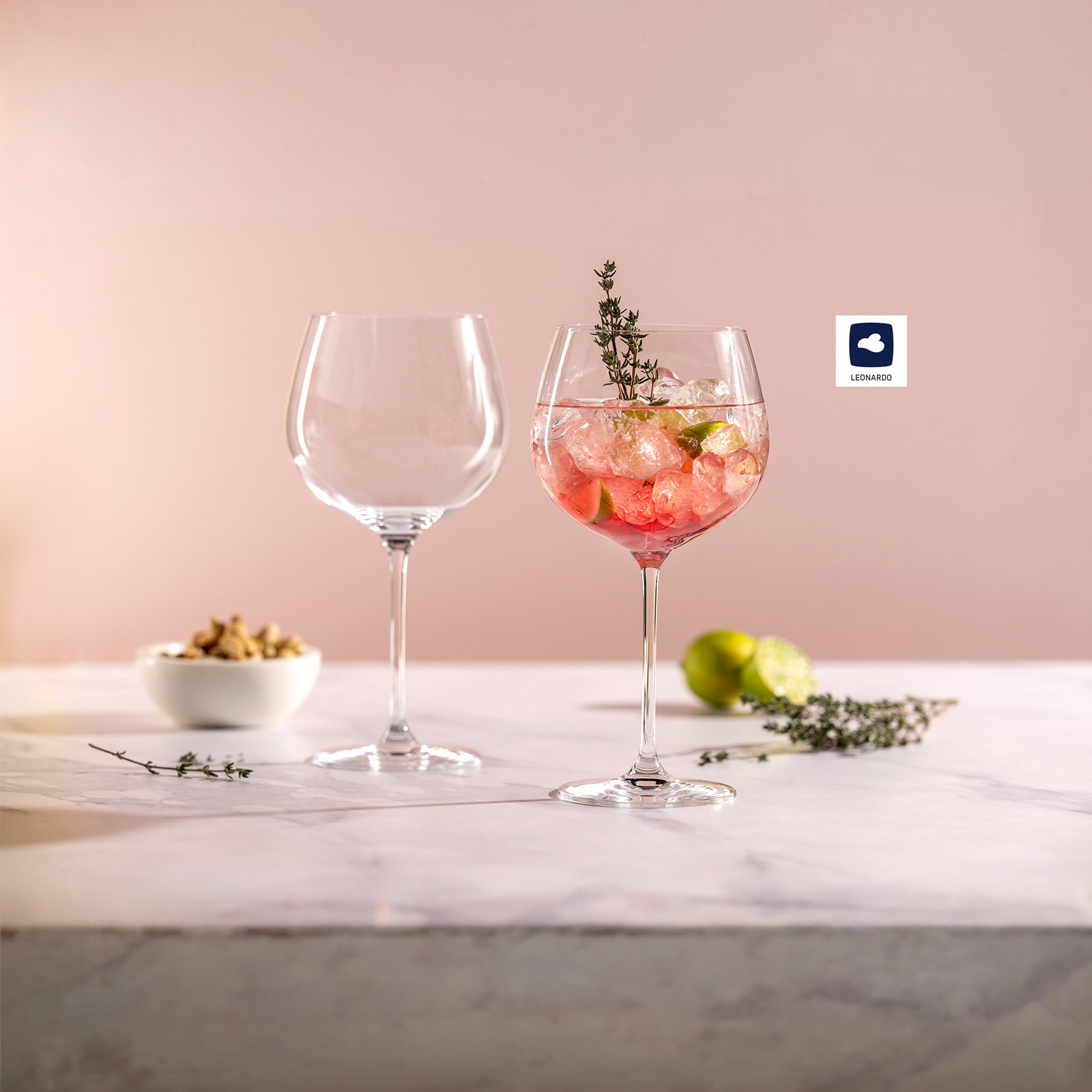 Zwei elegante Gin-Tonic-Gläser vor zartrosa Hintergrund, gefüllt mit rosasem Cocktail, Eiswürfeln, Limette und Thymian.