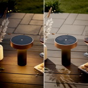 Runde, solarbetriebene Tischleuchte in Kupferoptik mit schwarzem Solarpanel oben, auf einem Holztisch im Außenbereich; Ansicht leicht von oben in schräger Perspektive.