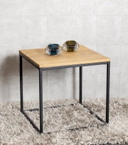Moderner quadratischer Beistelltisch mit heller Holzplatte und schwarzem Metallgestell auf flauschigem Teppich, mit zwei kleinen geriffelten Glasvasen obenauf; Perspektive: leicht schräg von vorne in Augenhöhe