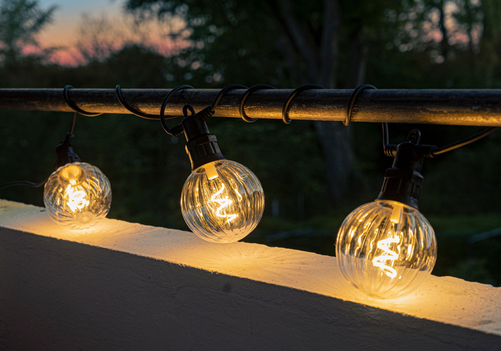 Lichterkette mit klaren, kugelförmigen LED-Lampen, die an einem Geländer bei Dämmerung aufgehängt sind.