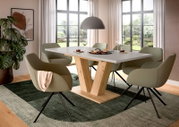 Moderner Esstisch mit weißer Tischplatte und V-förmigem Holzgestell, umgeben von sechs olivgrünen Polsterstühlen in einem hellen Esszimmer; Perspektive: leicht seitliche Frontansicht auf Tisch und Sitzgruppe