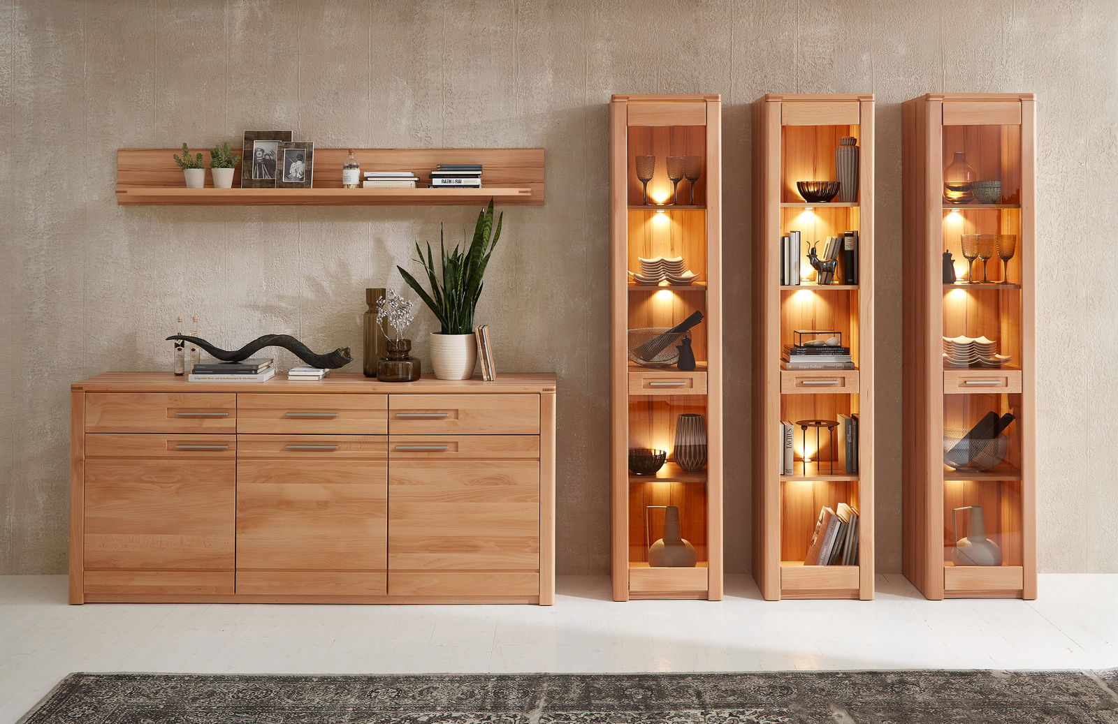 Schmales Regal aus Holz mit Glastüren, beleuchtet, frontal fotografiert, neben einer Kommode und einem Wandregal.