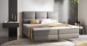 Modern boxspringbed met grijze bekleding en een hoog hoofdeinde, gezien vanaf de zijkant, gedecoreerd met kussens en een deken in neutrale tinten, in een stijlvolle slaapkamer.