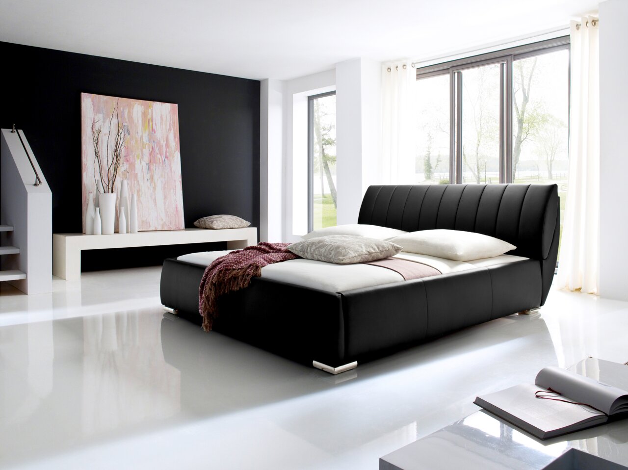 Modern gestoffeerd bed in zwart leer, gezien vanaf de zijkant, in een lichte kamer met grote ramen en een stijlvolle inrichting.