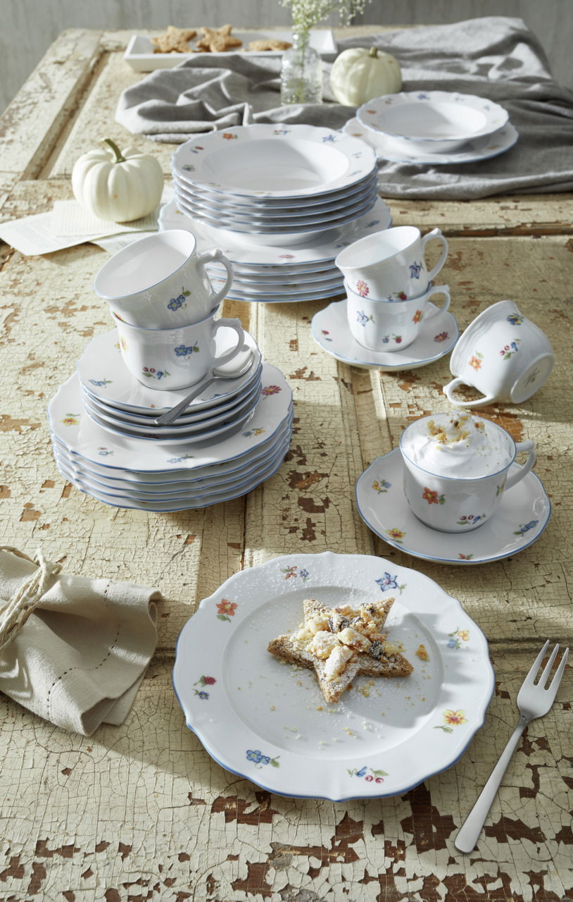Ein stilvolles Kombiservice-Set mit floralem Muster, bestehend aus Tellern, Tassen und Untertassen, auf einem rustikalen Holztisch arrangiert. Die Perspektive zeigt das Set von oben, mit einem dekorativen Sternkeks auf einem Teller im Vordergrund.