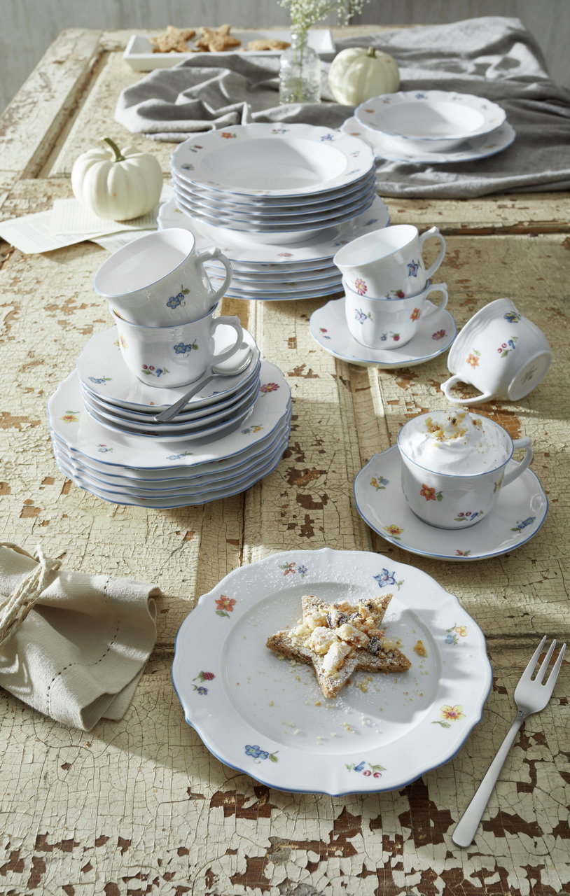 Ein stilvolles Kombiservice-Set mit floralem Muster, bestehend aus Tellern, Tassen und Untertassen, auf einem rustikalen Holztisch arrangiert. Die Perspektive zeigt das Set von oben, mit einem dekorativen Sternkeks auf einem Teller im Vordergrund.