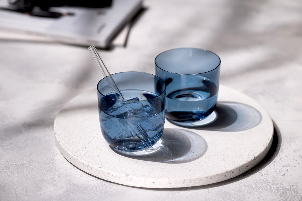 Zwei blaue Wassergläser auf einem runden, weißen Untersetzer, eines mit einem Glasstrohhalm und Eiswürfeln, aus der Vogelperspektive fotografiert.