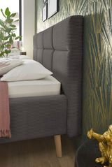 Seitliche Perspektive eines modernen Polsterbetts mit grauem, gestepptem Kopfteil und Holzbeinen, dekoriert mit Kissen und Decke.