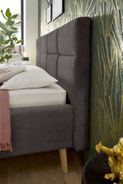 Zijaanzicht van een modern gestoffeerd bed met een grijs, doorgestikt hoofdeinde en houten poten, versierd met kussens en een deken.