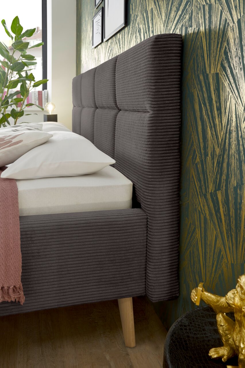 Zijaanzicht van een modern gestoffeerd bed met een grijs, doorgestikt hoofdeinde en houten poten, versierd met kussens en een deken.