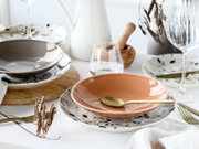 Stilvoll gedeckter Tisch mit terrakottafarbener Keramikschale auf gesprenkeltem Teller, goldfarbenem Löffel und Gläsern, aufgenommen aus leicht schräger Seitenperspektive (Nahaufnahme).