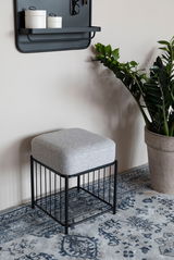 Moderner Hocker mit schwarzem Metallgestell und grauem Polster, seitliche Perspektive, auf einem gemusterten Teppich neben einer Pflanze.