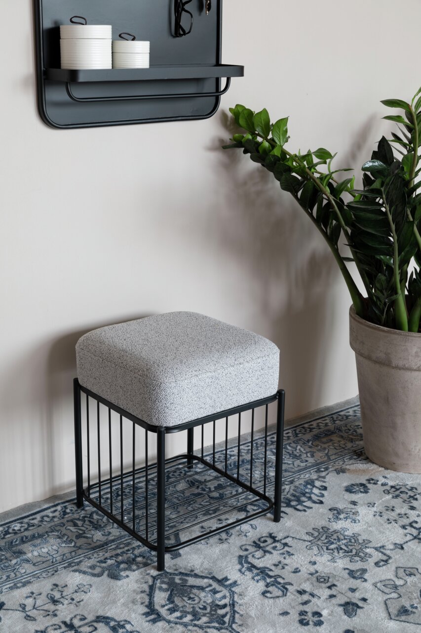 Moderne poef met een zwart metalen frame en grijze bekleding, gezien vanaf de zijkant, op een tapijt met patroon naast een plant.