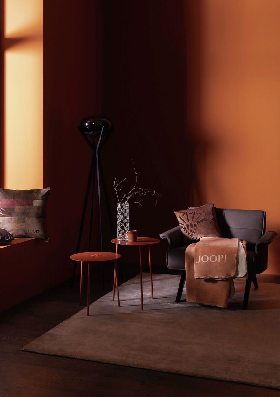 Gemütliche Wohnzimmeratmosphäre mit einem braunen Sessel, dekoriert mit einem Kissen und einer Decke von JOOP! in warmen Erdtönen, seitliche Perspektive.