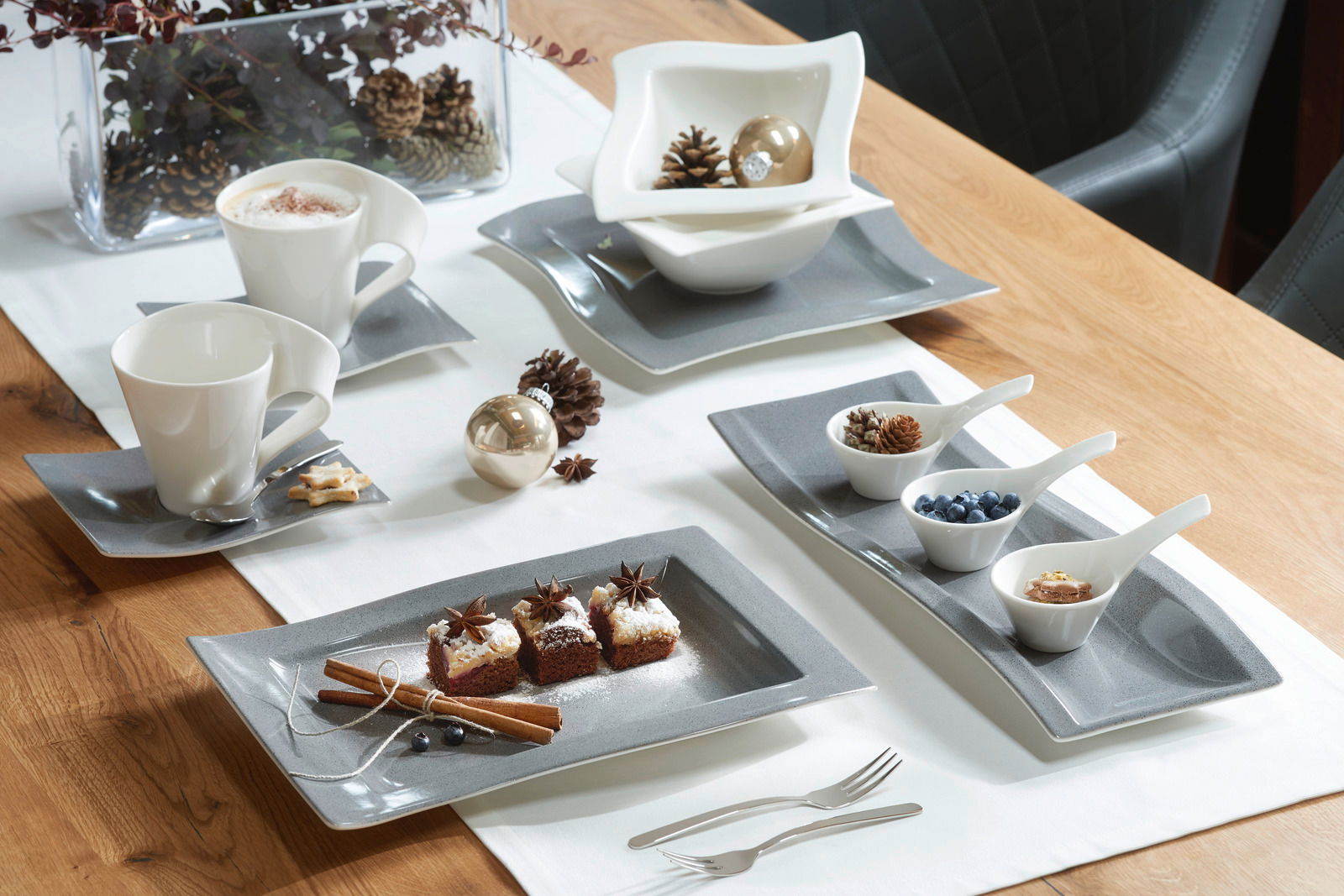 Elegantes Geschirrset auf einem Holztisch, bestehend aus weißen Tassen und Schalen auf grauen Tellern, dekoriert mit weihnachtlichen Elementen wie Tannenzapfen und Zimtstangen. Perspektive von oben.