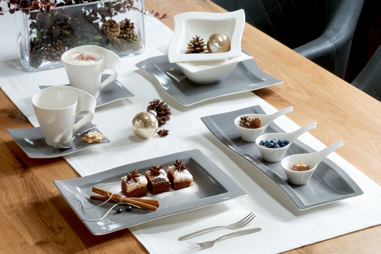 Elegantes Geschirrset auf einem Holztisch, bestehend aus weißen Tassen und Schalen auf grauen Tellern, dekoriert mit weihnachtlichen Elementen wie Tannenzapfen und Zimtstangen. Perspektive von oben.