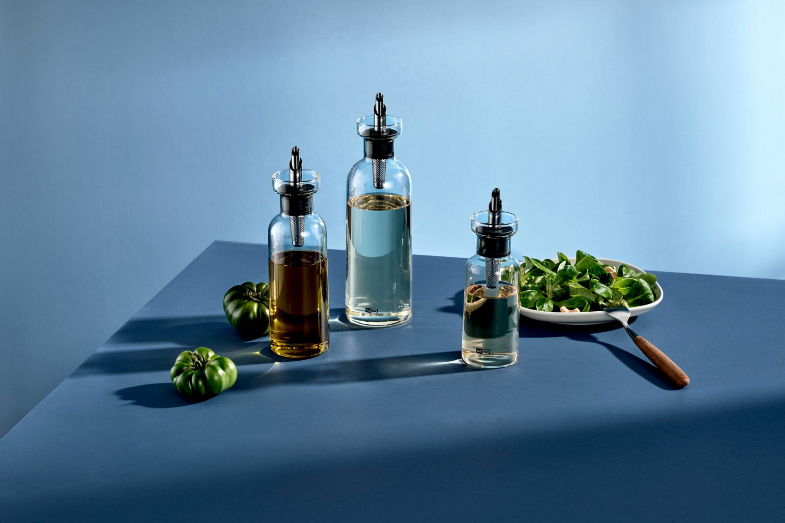 Drei transparente Glas-Öl- und Essigspender mit Ausgießer und integriertem Filter auf blauem Tisch neben Salatschüssel und grünen Tomaten, in leichter Schrägansicht von vorne