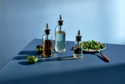 Drei transparente Glas-Öl- und Essigspender mit Ausgießer und integriertem Filter auf blauem Tisch neben Salatschüssel und grünen Tomaten, in leichter Schrägansicht von vorne