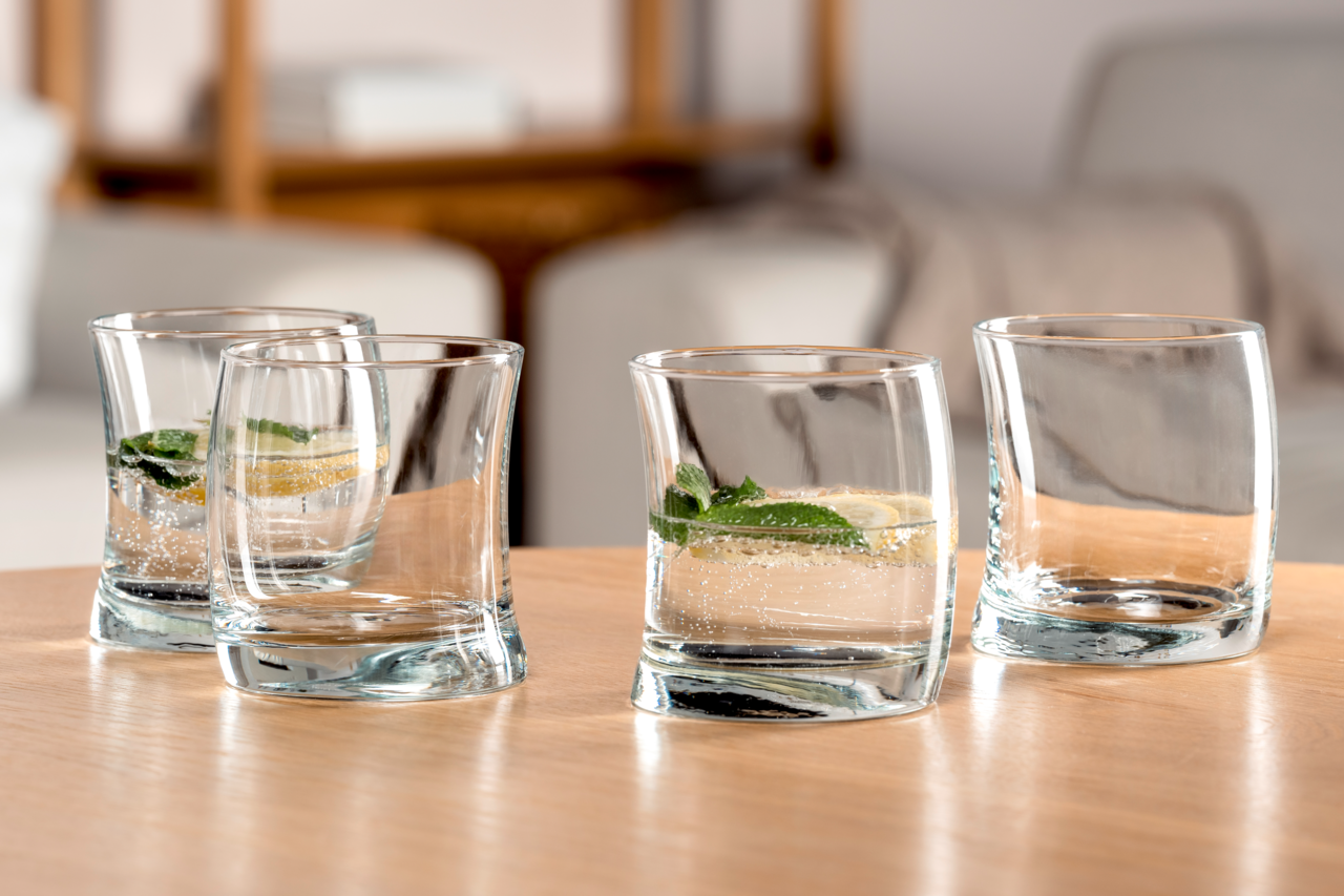 Vier elegante, leicht geschwungene Gläser auf einem Holztisch, zwei mit Getränk und Minzblättern gefüllt, aus seitlicher Perspektive fotografiert.