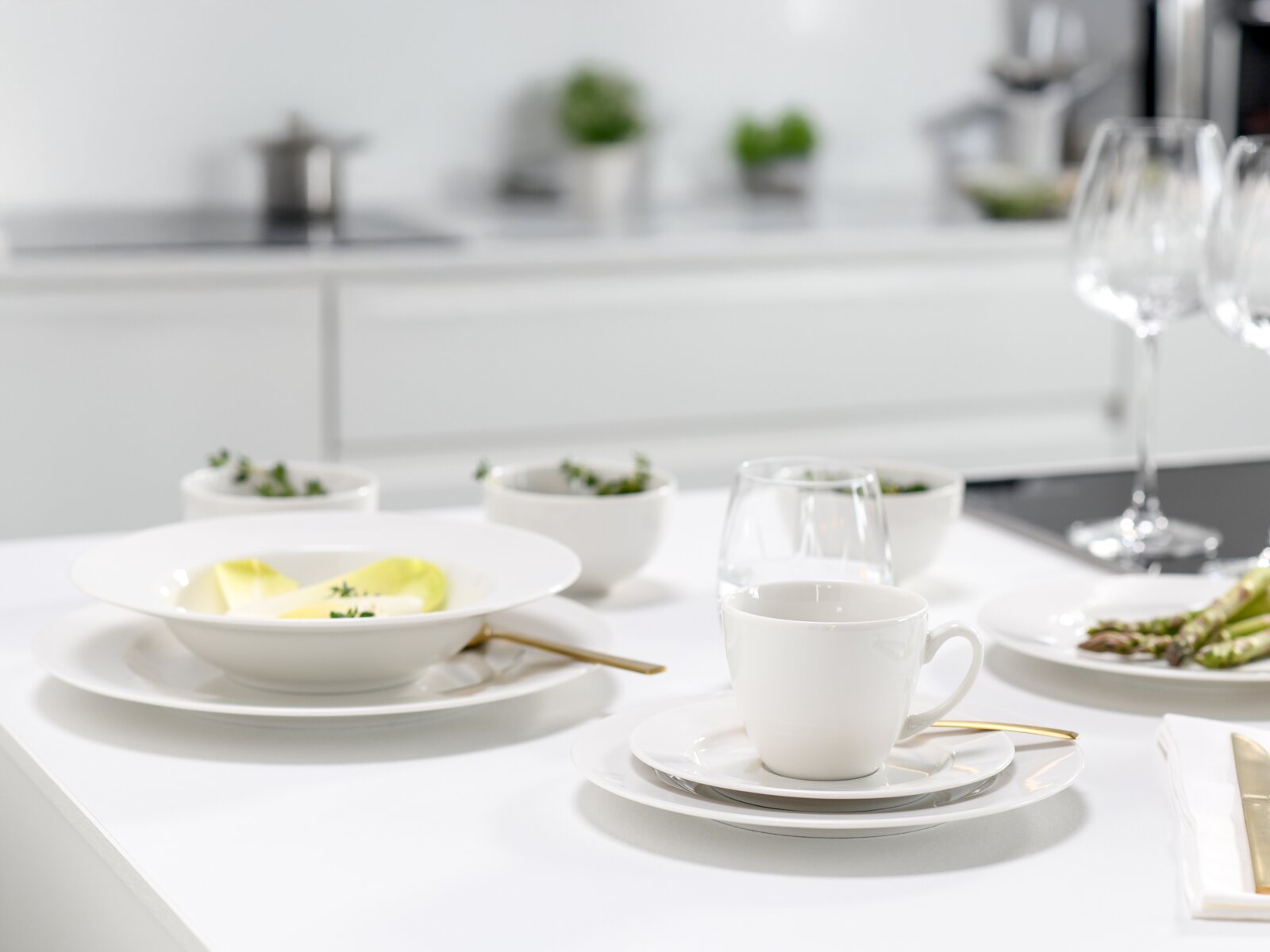 Modern gedeckter Esstisch mit weißem Porzellan-Geschirr, Kaffeetasse auf Untertasse und goldfarbenem Besteck in heller Küche, Perspektive: leicht seitlich von vorne auf Tischhöhe