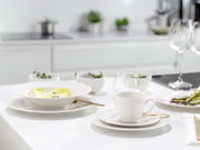 Modern gedeckter Esstisch mit weißem Porzellan-Geschirr, Kaffeetasse auf Untertasse und goldfarbenem Besteck in heller Küche, Perspektive: leicht seitlich von vorne auf Tischhöhe