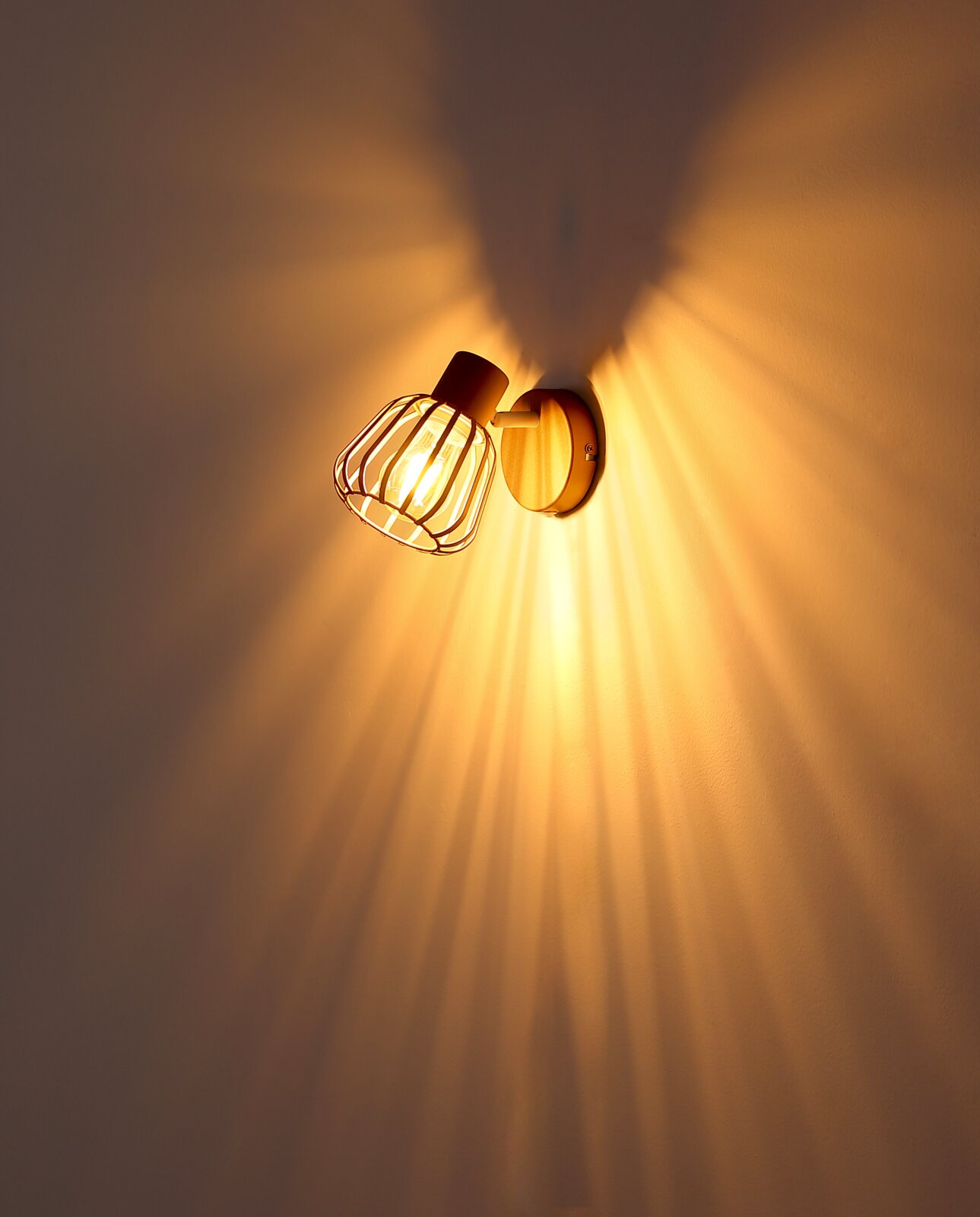 Eingeschaltete Wandlampe im Industrial-Design mit Metallgitter-Schirm und sichtbarer Glühbirne, warmes Licht mit Strahlenmuster an der Wand, fotografiert in leichter Untersicht aus schräger Seitenperspektive.