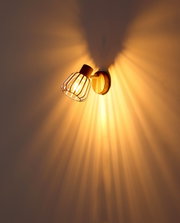 Eingeschaltete Wandlampe im Industrial-Design mit Metallgitter-Schirm und sichtbarer Glühbirne, warmes Licht mit Strahlenmuster an der Wand, fotografiert in leichter Untersicht aus schräger Seitenperspektive.