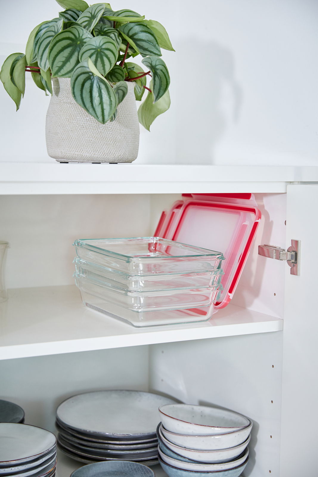 Rechteckige Glasbehälter mit roten Deckeln, gestapelt in einem weißen Küchenschrank, seitliche Perspektive