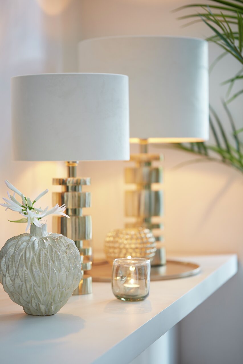 Zwei elegante Tischlampen mit zylindrischen, weißen Lampenschirmen und goldenen Sockeln auf einem Tisch, seitliche Perspektive.