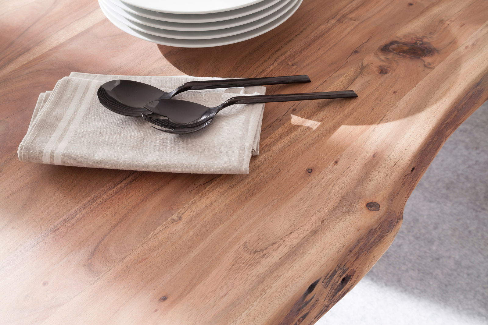 Nahaufnahme eines Esstisches aus Holz mit natürlicher Maserung, darauf liegen ein Stapel weißer Teller, ein Stoffserviette und zwei schwarze Löffel. Perspektive von oben.