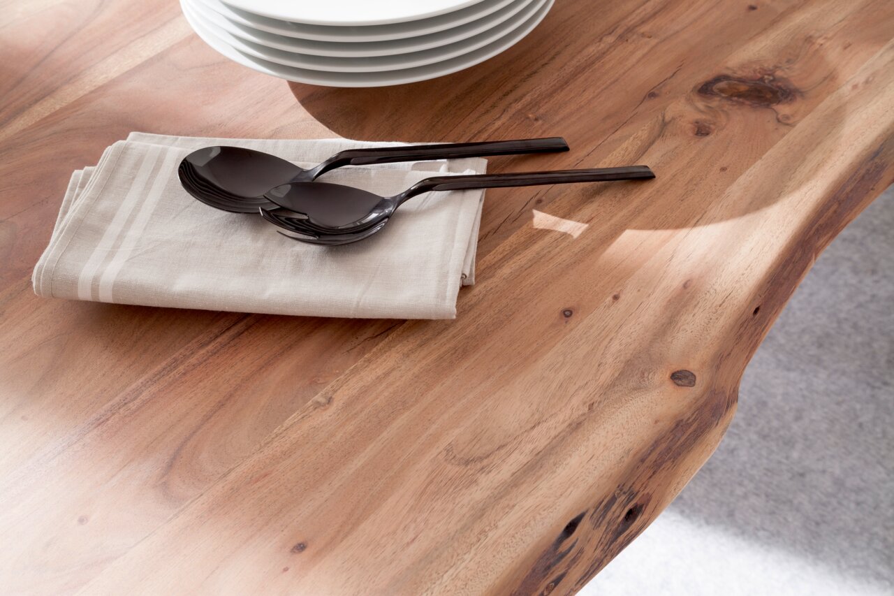 Nahaufnahme eines Esstisches aus Holz mit natürlicher Maserung, darauf liegen ein Stapel weißer Teller, ein Stoffserviette und zwei schwarze Löffel. Perspektive von oben.