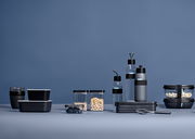 Verschiedene schwarze und transparente Lunchboxen und Flaschen in einer modernen, minimalistischen Anordnung auf einem blauen Hintergrund, seitliche Perspektive.