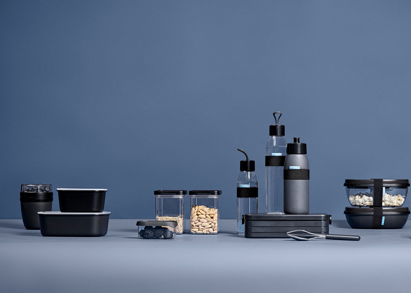 Verschiedene schwarze und transparente Lunchboxen und Flaschen in einer modernen, minimalistischen Anordnung auf einem blauen Hintergrund, seitliche Perspektive.