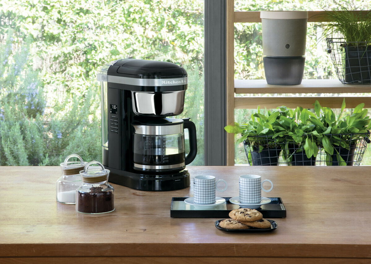 Kaffeemaschine in onyx schwarz auf einem Holztisch, umgeben von Kaffeetassen, Keksen und Pflanzen im Hintergrund, seitliche Perspektive.