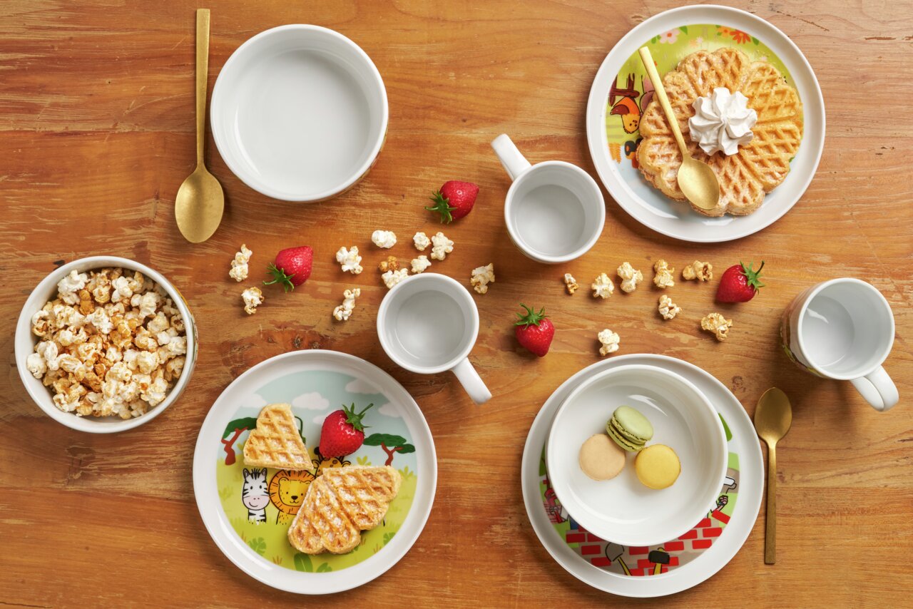 Vogelperspectief op een kinderservies met bouwplaatsmotief, bestaande uit borden, kommen en kopjes, versierd met wafels, popcorn en aardbeien.