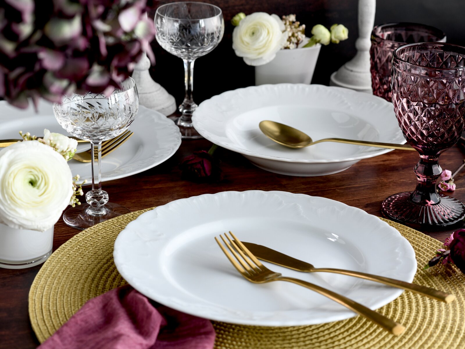 Festlich gedeckter Holztisch mit weißen Porzellantellern und -schalen, goldfarbenem Besteck sowie Kristall- und roséfarbenen Gläsern, dekoriert mit weißen Blumen; Perspektive: leicht erhöhte schräge Ansicht von vorne.
