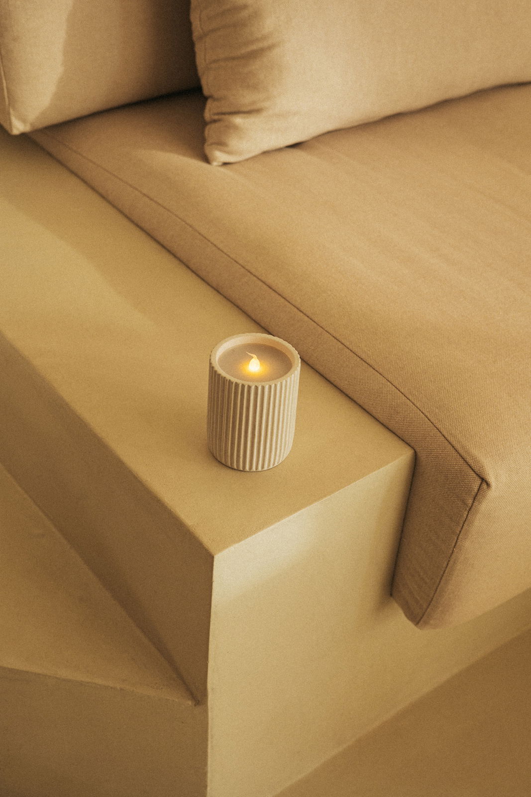 Outdoor-Kerze in einem geriffelten, beigen Behälter, auf einer beigen Treppenstufe neben einem Sofa platziert, aus einer leicht erhöhten Perspektive fotografiert.