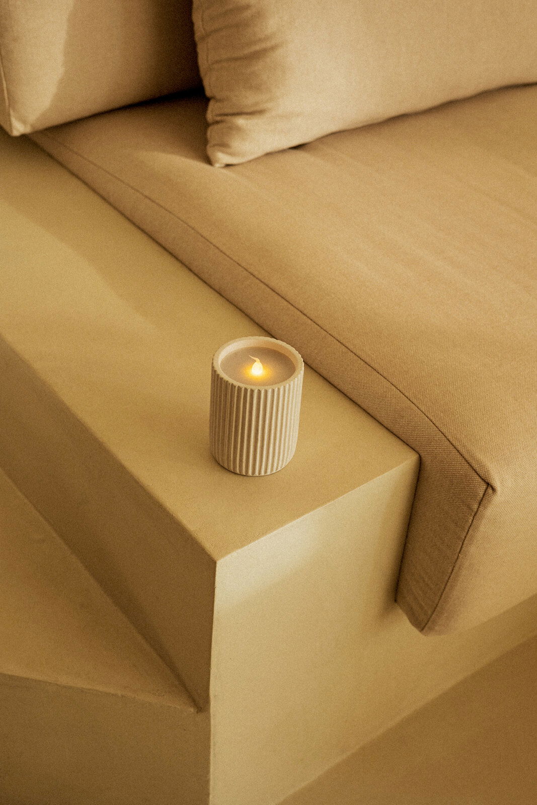 Outdoor-Kerze in einem geriffelten, beigen Behälter, auf einer beigen Treppenstufe neben einem Sofa platziert, aus einer leicht erhöhten Perspektive fotografiert.