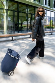 Een vrouw trekt een donkerblauwe boodschappentrolley met twee wielen en een uitschuifbaar handvat over een trottoir voor een modern glazen gebouw; zijaanzicht vanuit een licht onderhoek.