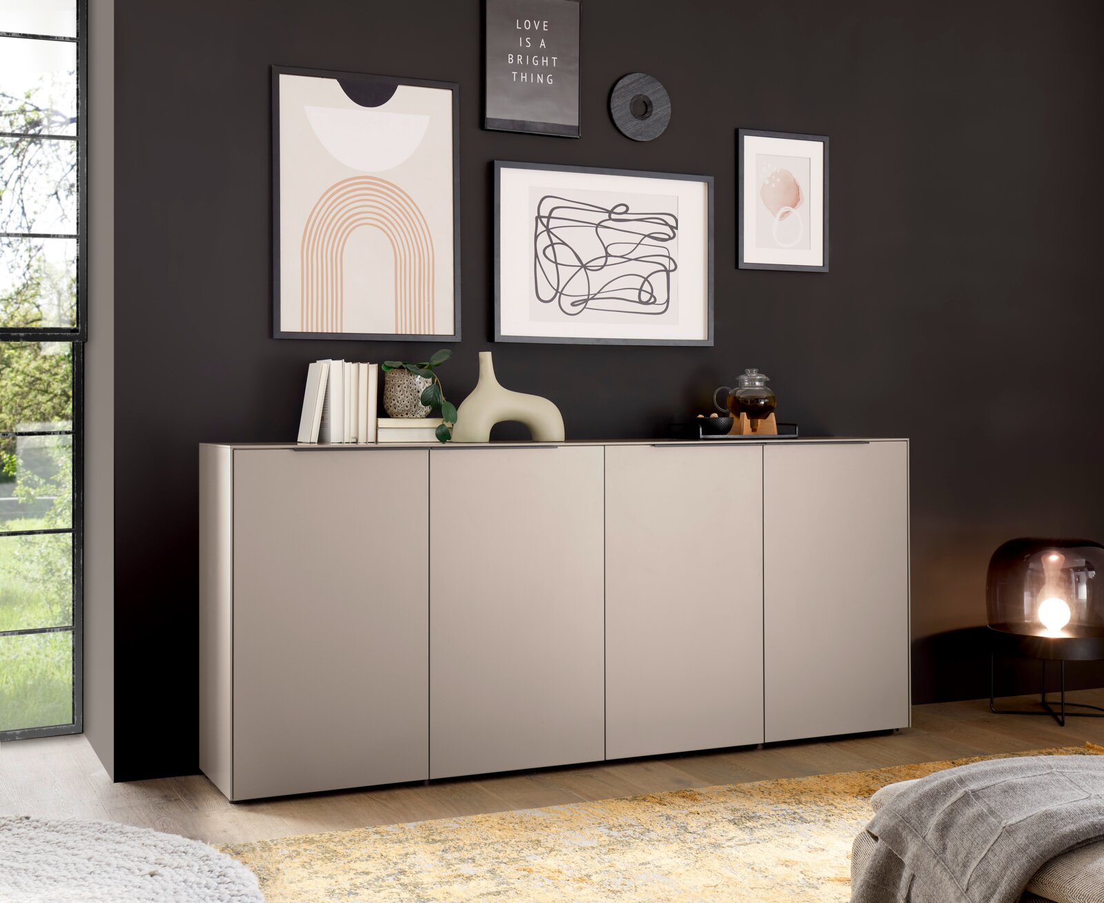 Modernes, niedriges Sideboard in Hellgrau mit vier Fronttüren vor dunkler Wand mit gerahmten Kunstdrucken; dekoriert mit Büchern, Vase und Teeset, aufgenommen in frontaler Perspektive leicht von links.
