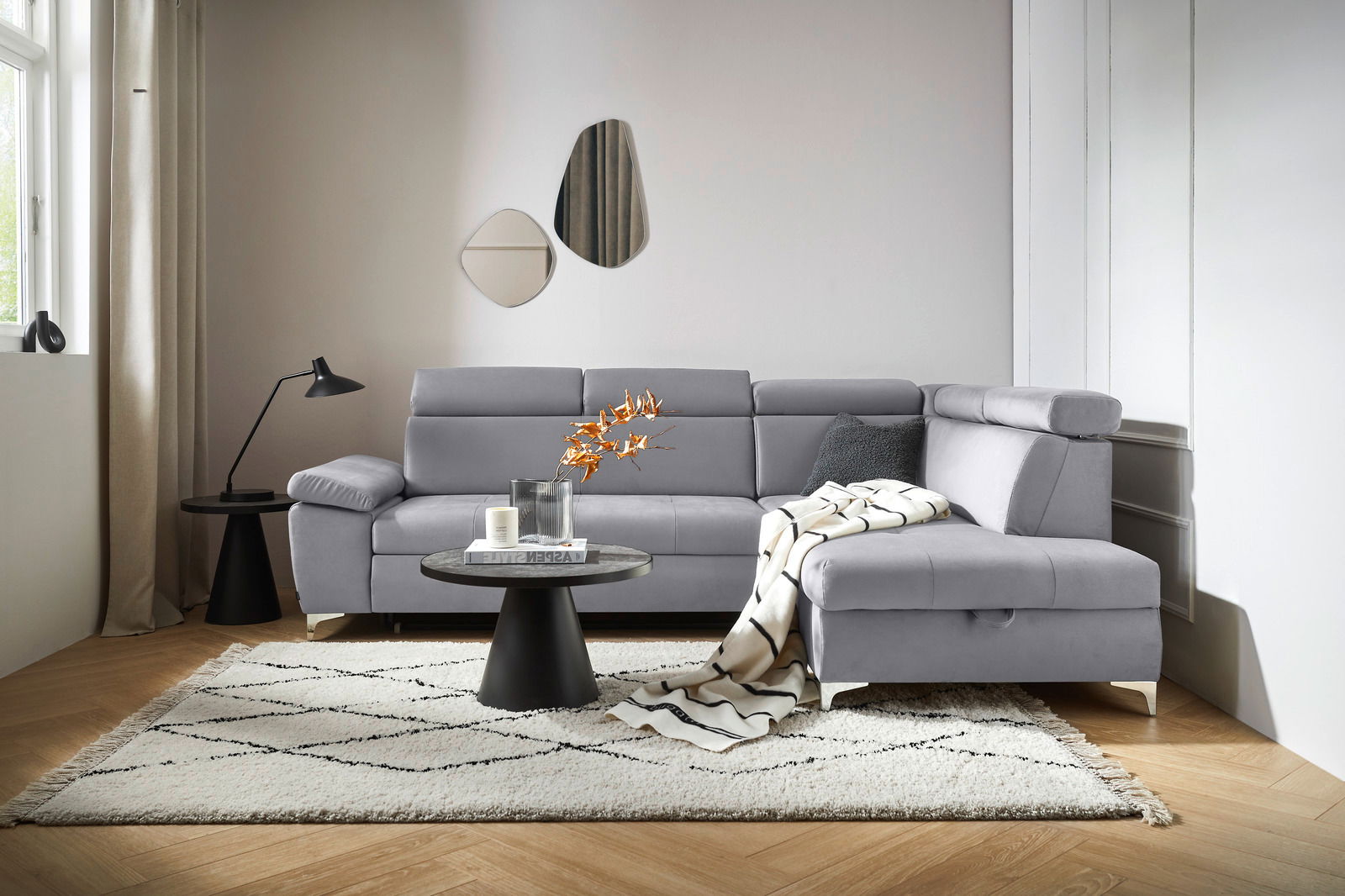 Graue Rundecke (L) in einem modernen Wohnzimmer, von vorne betrachtet, mit einem Beistelltisch, einer Lampe und einem Teppich.