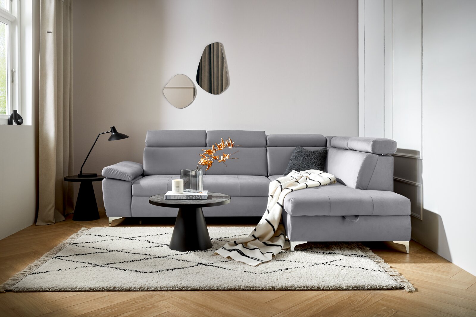 Grijze hoekbank (L) in een moderne woonkamer, gezien vanaf de voorkant, met een bijzettafeltje, een lamp en een vloerkleed.