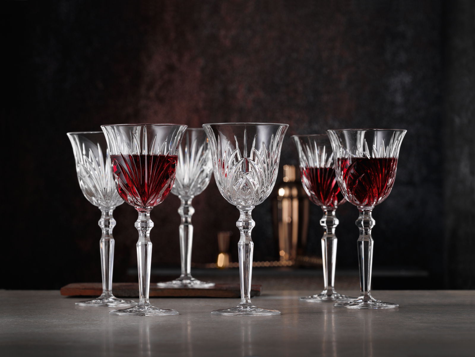 Sechs elegante Rotweinkelche aus Kristallglas, teilweise gefüllt mit Rotwein, auf einem Tisch vor dunklem Hintergrund, seitliche Perspektive.