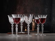 Sechs elegante Rotweinkelche aus Kristallglas, teilweise gefüllt mit Rotwein, auf einem Tisch vor dunklem Hintergrund, seitliche Perspektive.