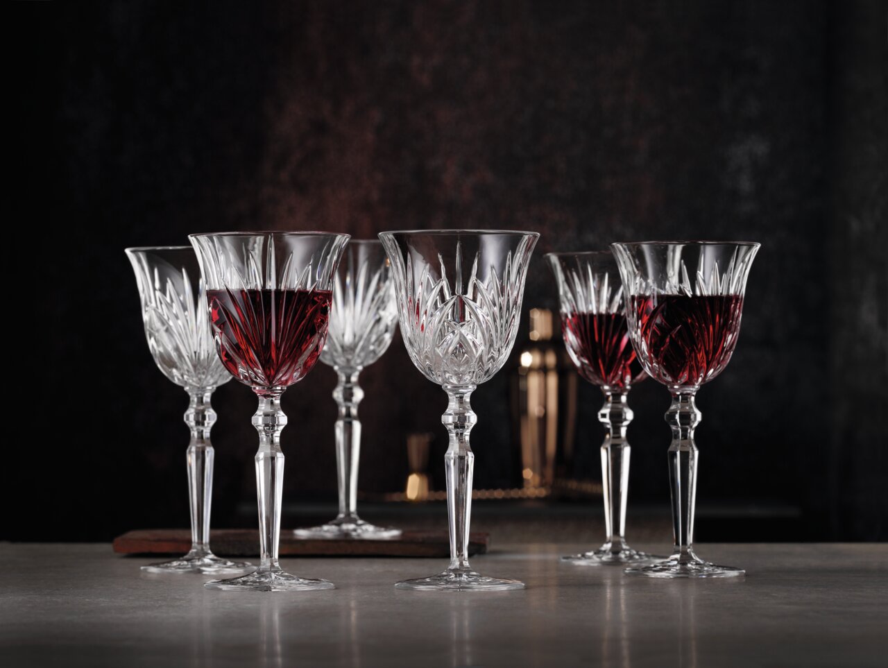 Sechs elegante Rotweinkelche aus Kristallglas, teilweise gefüllt mit Rotwein, auf einem Tisch vor dunklem Hintergrund, seitliche Perspektive.