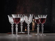 Sechs elegante Rotweinkelche aus Kristallglas, teilweise gefüllt mit Rotwein, auf einem Tisch vor dunklem Hintergrund, seitliche Perspektive.