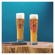 Zwei Weizenbiergläser mit Schaumkrone, eines mit einem Tiger-Design und das andere mit einem Löwen-Design, auf einem Holztisch vor blauem Hintergrund, seitliche Perspektive.