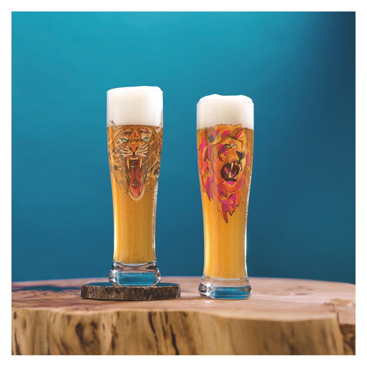 Zwei Weizenbiergläser mit Schaumkrone, eines mit einem Tiger-Design und das andere mit einem Löwen-Design, auf einem Holztisch vor blauem Hintergrund, seitliche Perspektive.