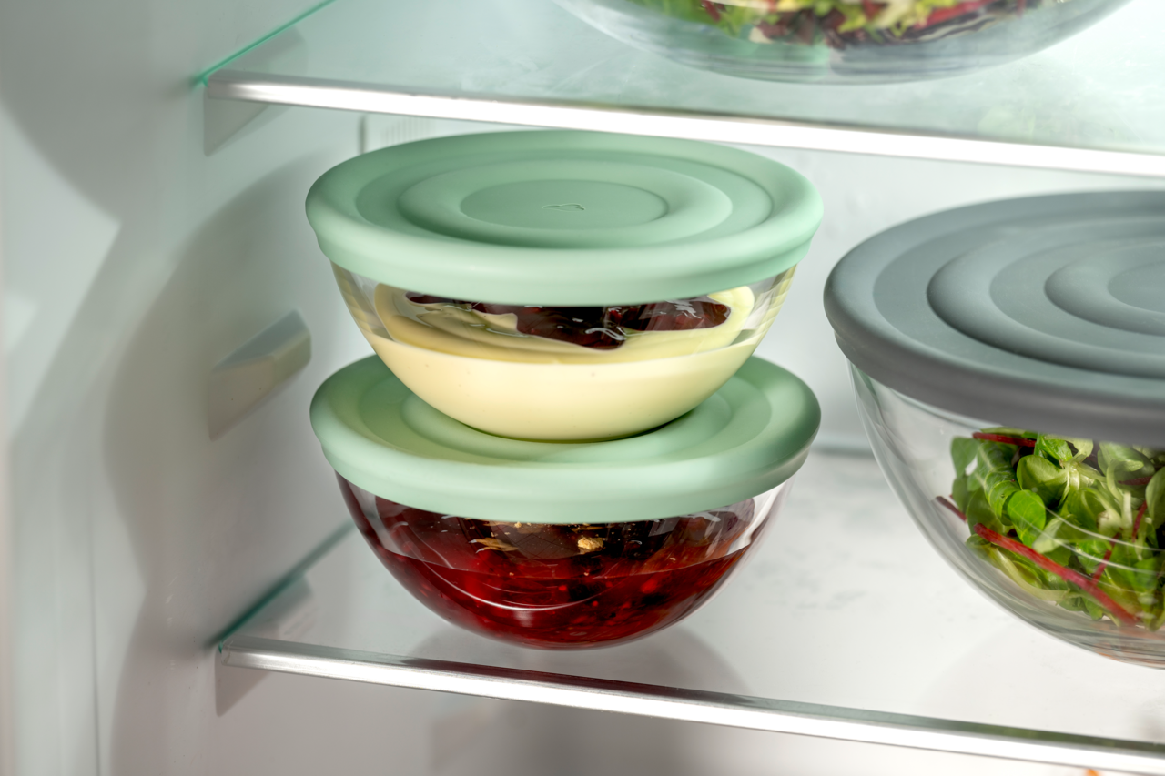 Zwei stapelbare Glasschalen mit grünen Deckeln, gefüllt mit Dessert und Kompott, in einem Kühlschrank von der Seite betrachtet.
